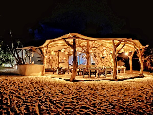 An outdoor wooden structure with tables and chairs, surrounded by sand.