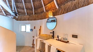 A bathroom with a thatched roof, a round mirror, a sink with a beige countertop, and a window. - Hotel Qamar Lux (Tulum)