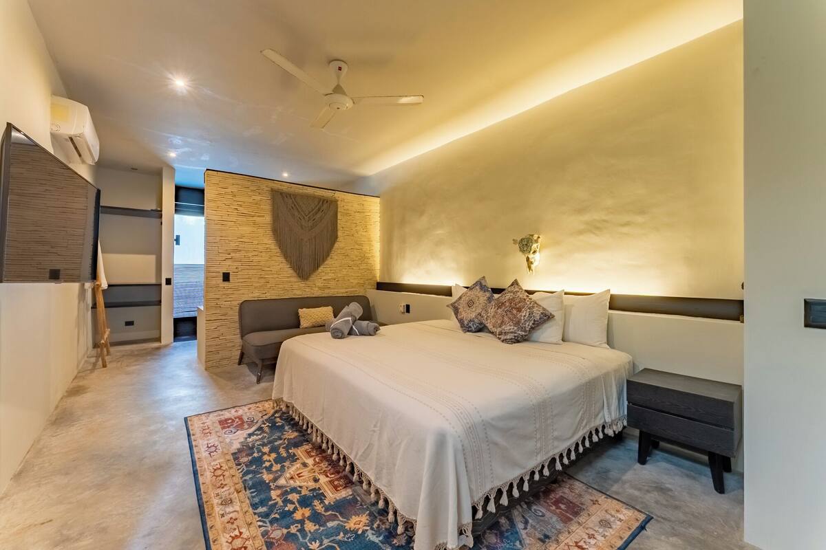 Une chambre moderne avec un grand lit, une tête de lit dotée d’un luminaire, un ventilateur de plafond et un tapis à motifs.