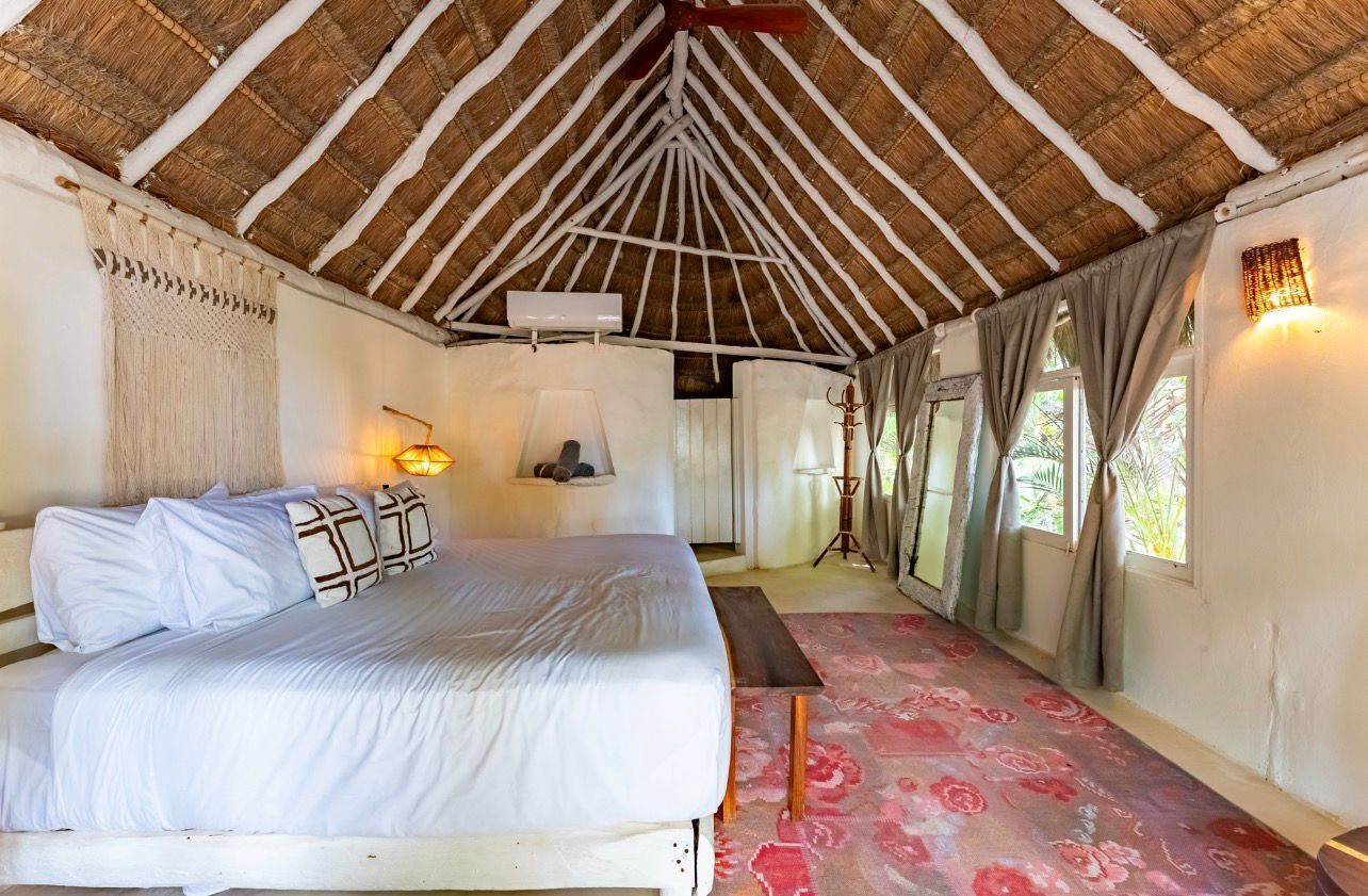A room with a bed, a bench, a lamp, a wall-mounted light, a window with curtains, and a thatched roof.