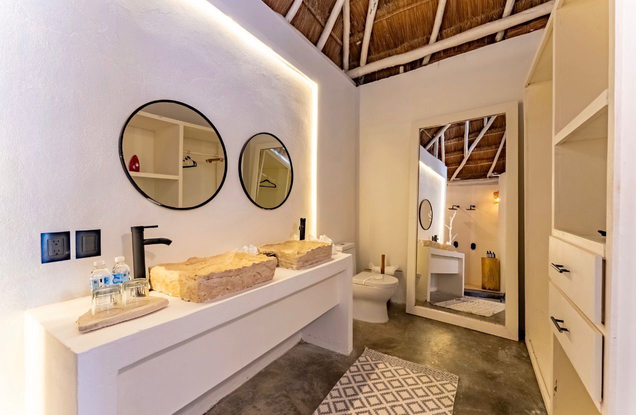 Un baño con un tocador blanco, dos espejos redondos, un lavamanos grande color beige, un inodoro y un mueble blanco pequeño.