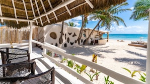A beachside resort with a thatched roof structure, white walls with circular cutouts, and a sandy beach with palm trees. - Hotel Qamar Lux (Tulum)