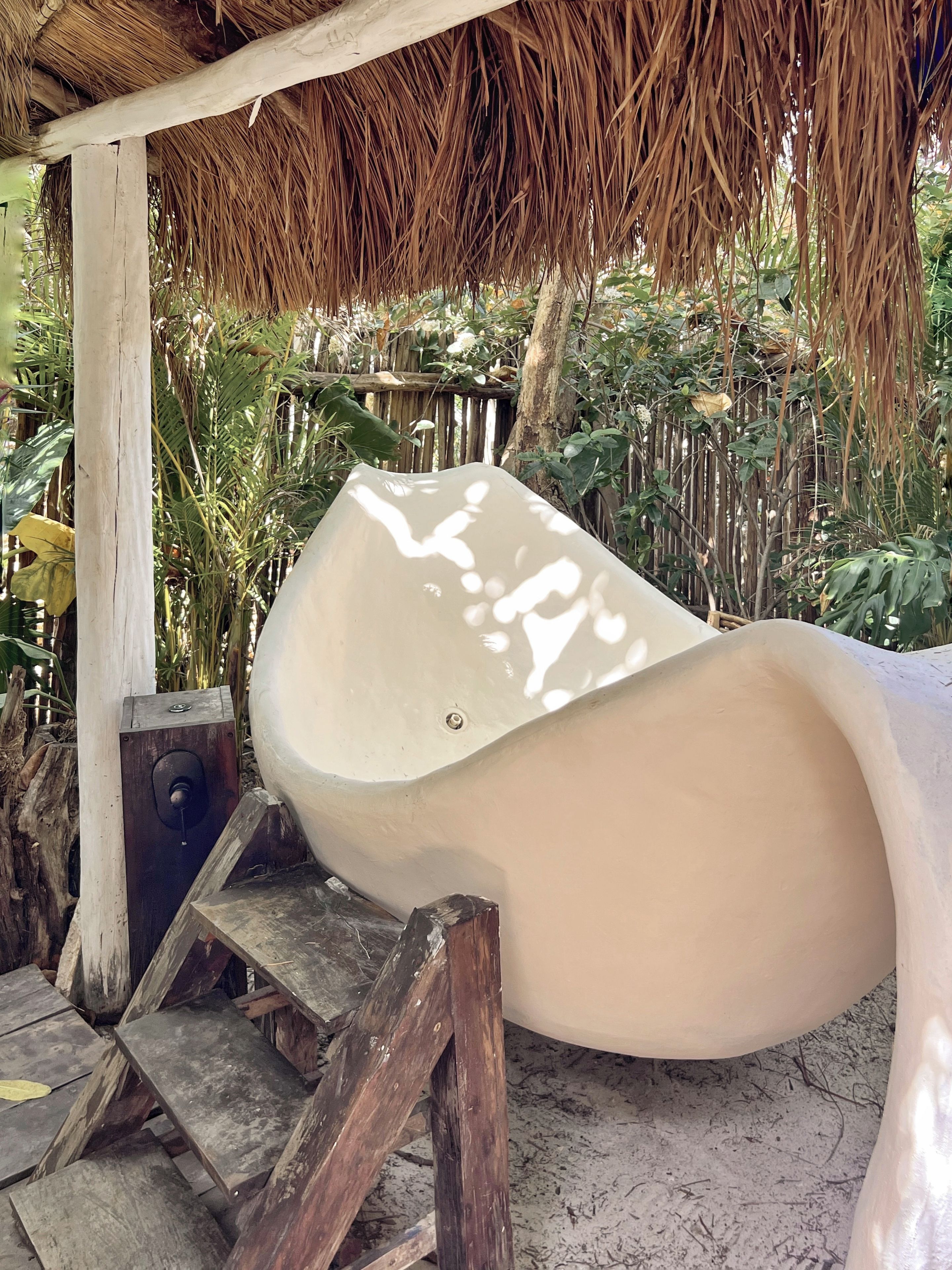 A white slide under a thatched roof structure with a wooden staircase leading up to it.