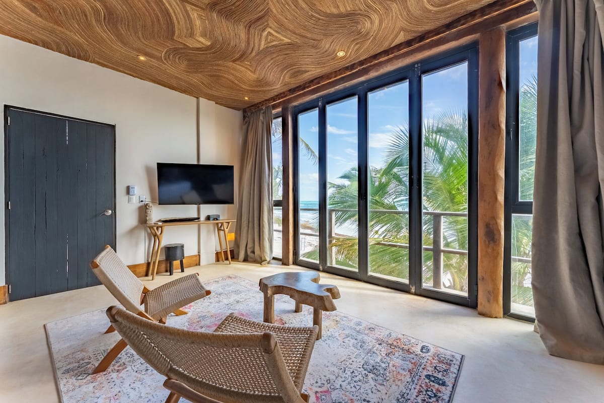 Une pièce dotée d’un grand téléviseur, de fauteuils en osier, d’un plafond en bois et offrant une vue sur la plage.