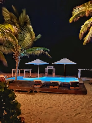 A nighttime poolside area with lounge chairs and umbrellas, a palm tree, and a clear pool. - Hotel Qamar Lux (Tulum)