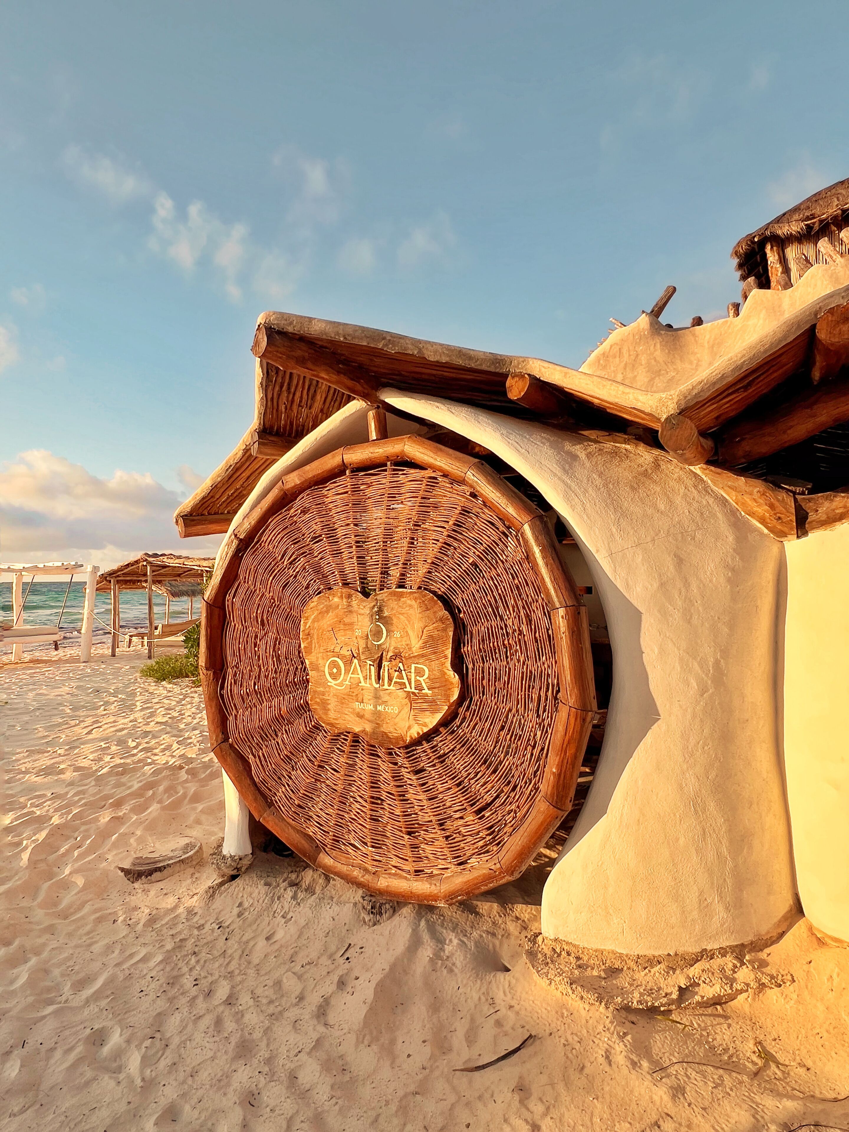 A beachside building with a woven circular structure labeled 'QAIJAR ITALY MEXICO' and a thatched roof.