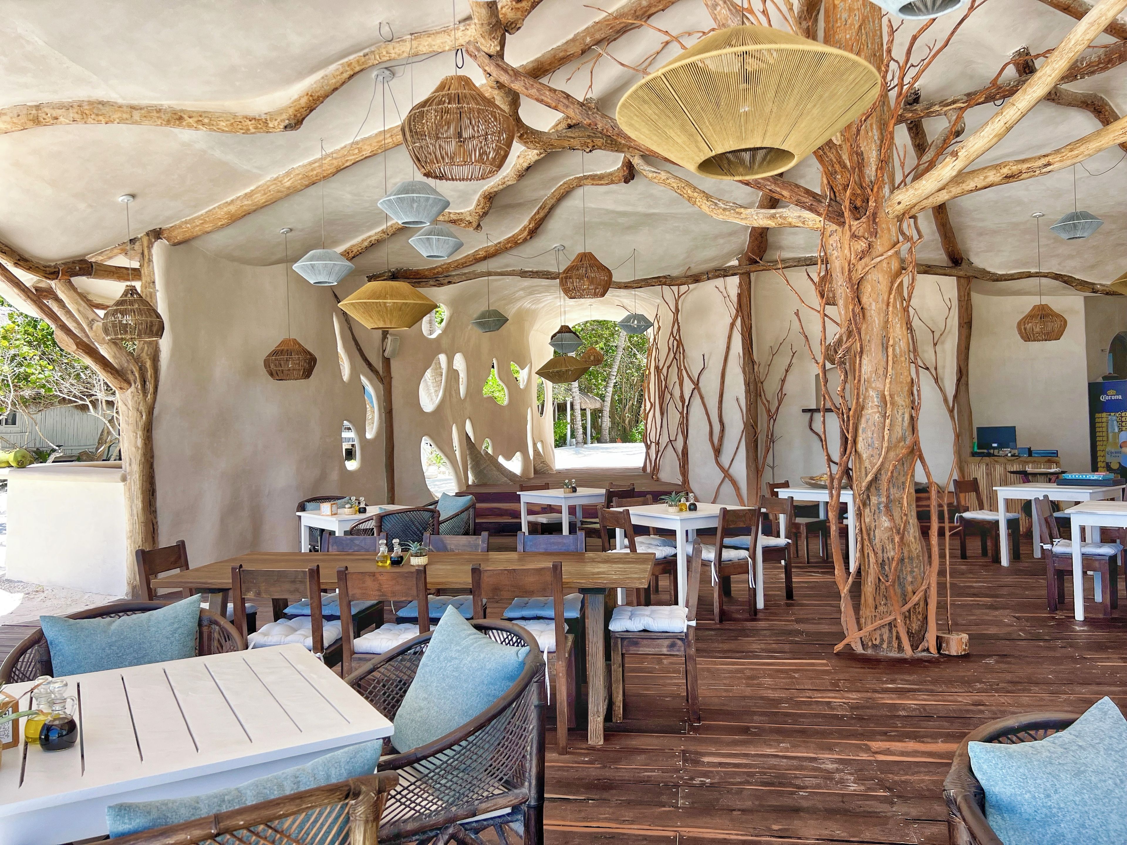 Een openluchtrestaurant met houten tafels en stoelen, hangende lampen en een boomstam als middelpunt.