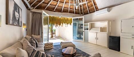 A modern living room with a thatched ceiling, a sofa set, a small round table, and a kitchen area visible through an open door.