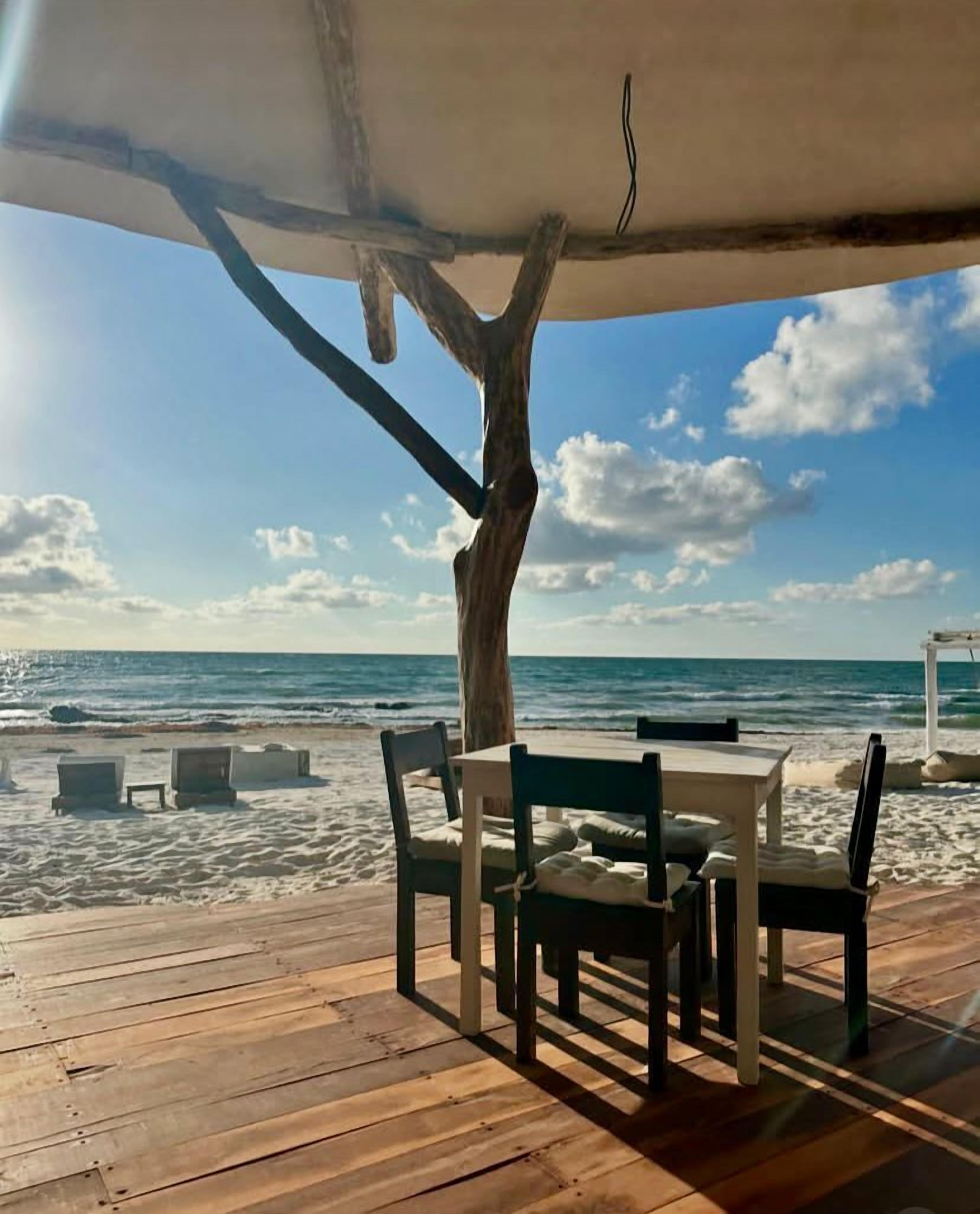 A wooden deck with a table and chairs, overlooking a beach with the ocean and sky.