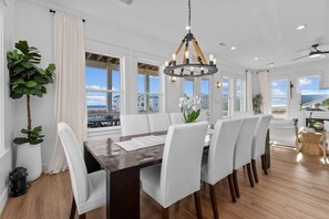 Un comedor con una larga mesa, sillas blancas, una araña y ventanas amplias con vistas.