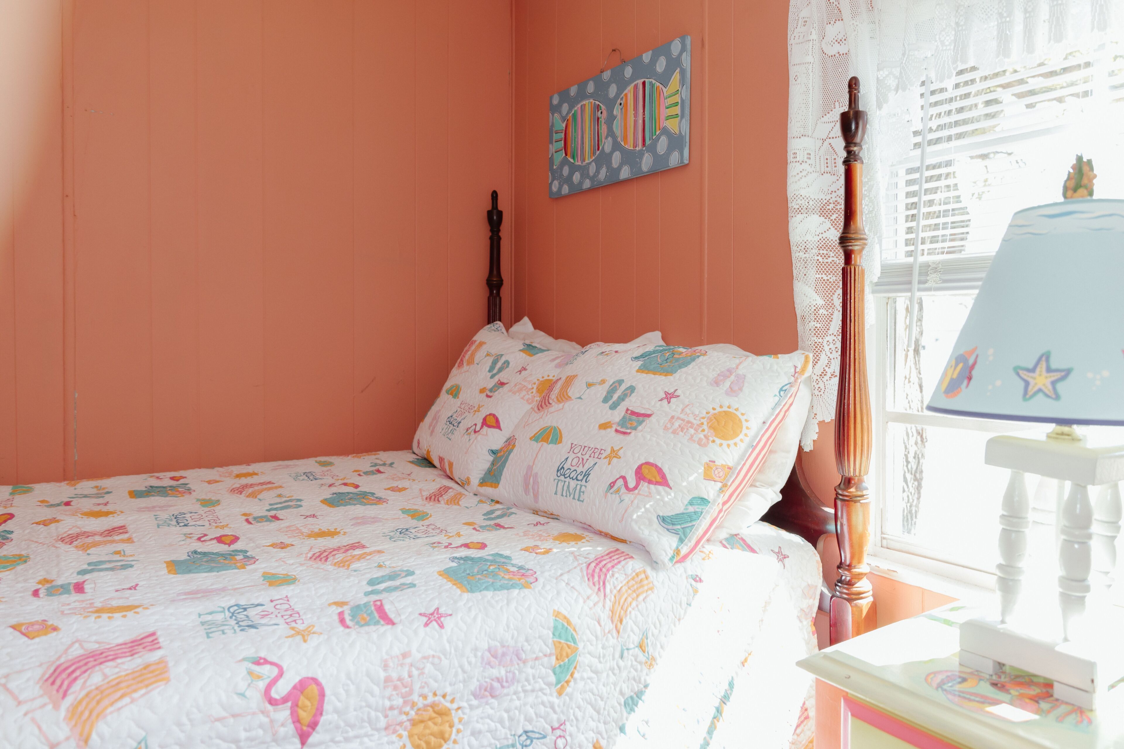 Une chambre à coucher comprenant un lit, une table de chevet, une lampe, une fenêtre avec des rideaux en dentelle et un mur orné d’une œuvre d’art sur le thème des poissons.