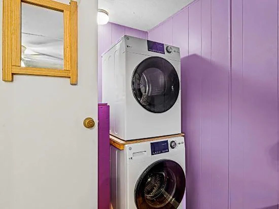 A laundry room with two stacked washing machines, a wooden-framed mirror, and purple walls.