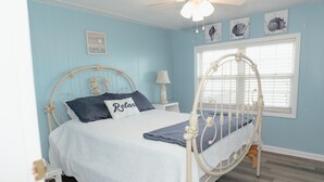 Een slaapkamer met een bed, nachtkastje, lamp, raam met jaloezieën en wanddecoratie.