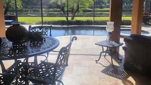 An outdoor patio with round tables and chairs, a view of a garden with palm trees, and a pond. - Charming 3-bedroom pool house in Sarasota, 5-minutes from Siesta Key beach (Sarasota)