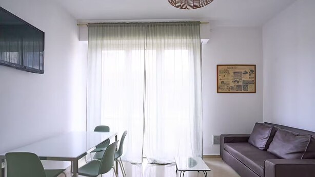 Een moderne woonkamer met een glazen eettafel, groene stoelen, een bruine bank en een groot raam met dunne gordijnen.