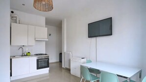 Een moderne keuken met witte kastjes, een fornuis en een spoelbak. Er is een eettafel met stoelen en een flatscreen televisie aan de muur bevestigd.