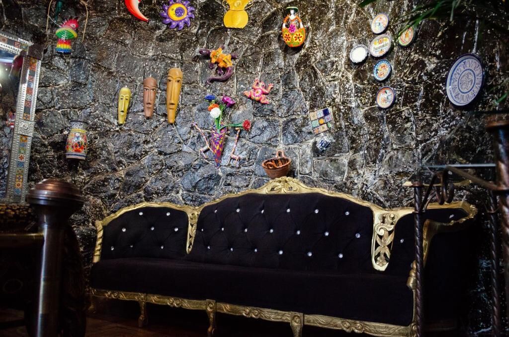 Una habitación con un sofá tapizado en negro con detalles dorados, una pared de piedra decorada con diversos objetos, y una botella de vino a la izquierda.