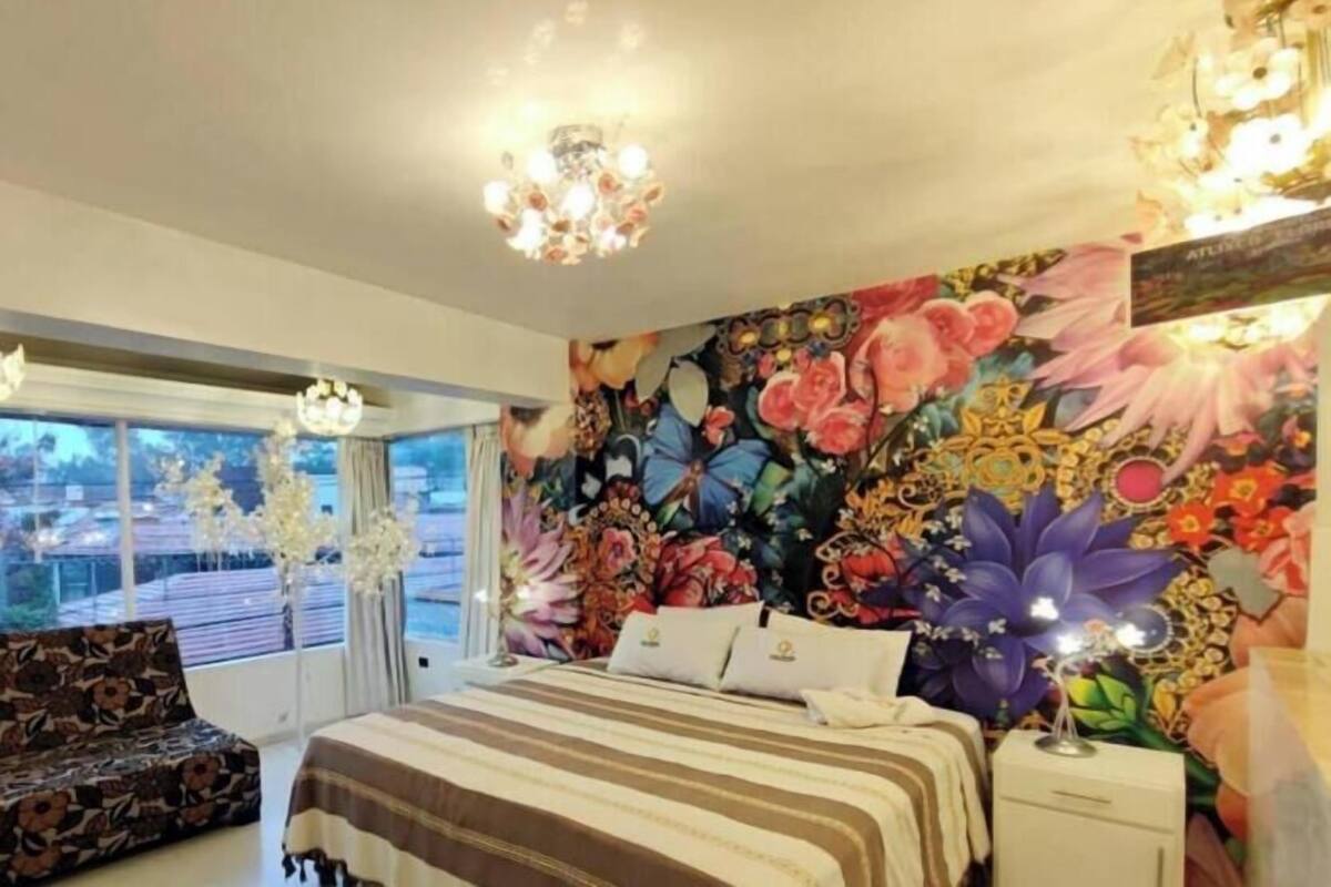 A bedroom with a floral mural, a bed with striped bedding, a sofa, and two chandeliers.
