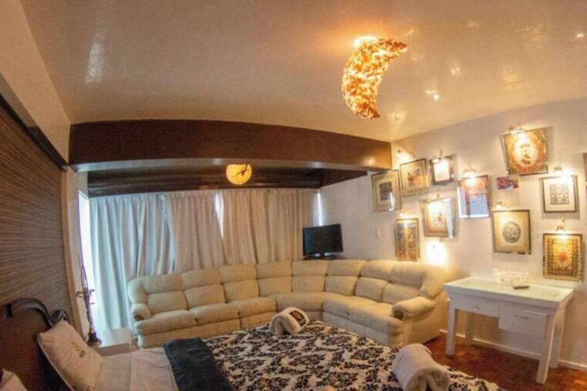 A hotel room with a large beige sofa, a small white table, and a television. The room has a ceiling light fixture and wall-mounted artwork.