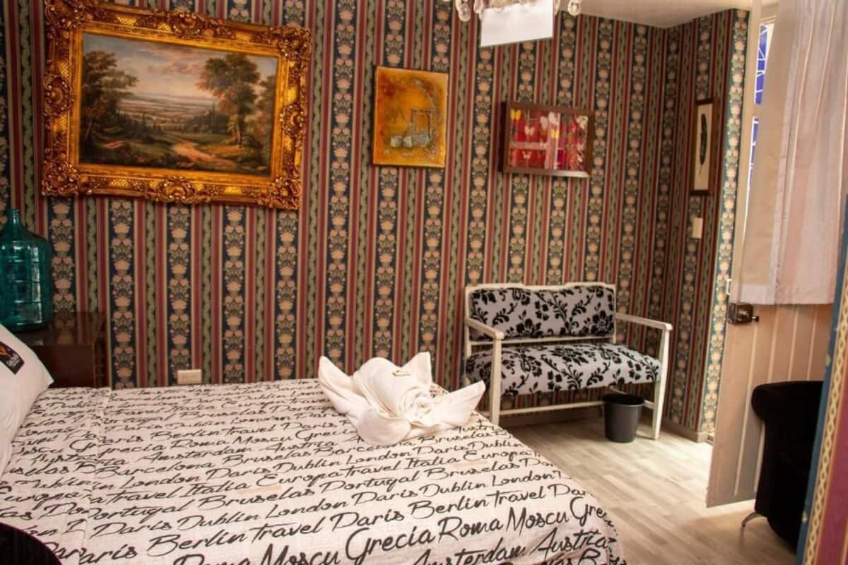 A hotel room with striped wallpaper, a bed with a travel-themed bedspread, a white chair, and a framed painting on the wall.