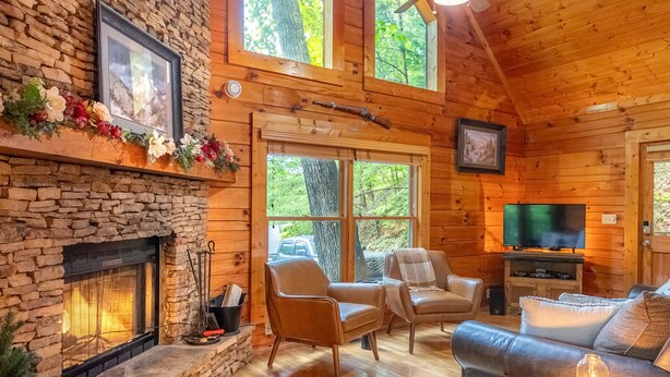 L’intérieur d’une cabane en rondins, avec une cheminée en pierre, un plancher en bois, un ventilateur de plafond, un téléviseur et un coin salon confortable.