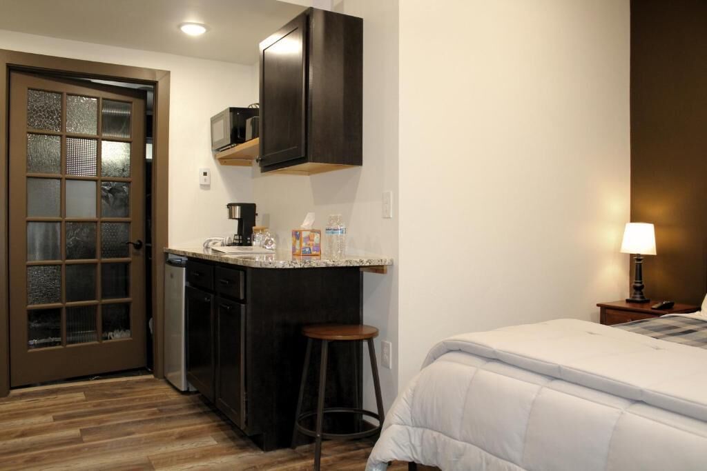 A compact hotel room with a kitchenette, a bed, a small stool, and a nightstand with a lamp.