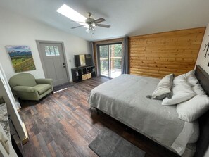 A bedroom with a bed, a chair, a TV, a wooden headboard, and a view through a glass door. - Two Bridges Lodge (Bailey)