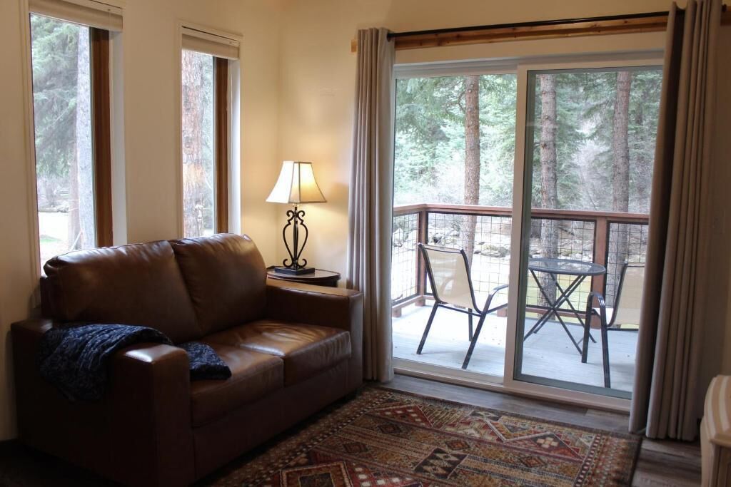 A living room with a brown leather sofa, a patterned rug, a table lamp, and a sliding glass door leading to an outdoor patio with chairs.