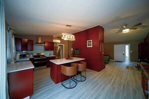 A modern kitchen with red cabinetry, a central island with stools, a ceiling fan, and a view of the living area. - Vino Villa | Cozy Escape near Wineries & Lake (Coloma)