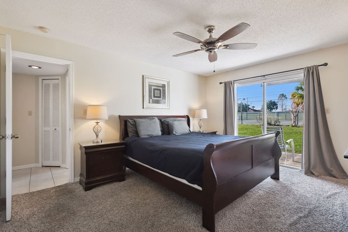 A bedroom with a bed, two bedside tables with lamps, a ceiling fan, a door, and a large window with a view of the outdoors.