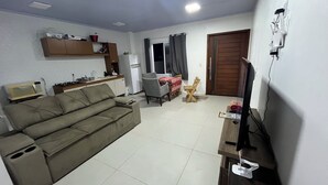 A living room with a sofa, a dining table with chairs, a TV, and a kitchen area with a refrigerator and microwave. - Semi-detached house in Praia Pérola (1 block from the beach) (Balneário Rosa do Mar)