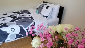 Un dormitorio con una cama cubierta con una colcha de diseño blanco y negro, un almohadón blanco y una mesita de noche con un jarrón de flores rosadas y una rosa blanca.
