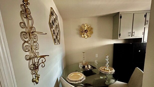 A dining area with a glass table set for two, white chairs, and decorative wall art.