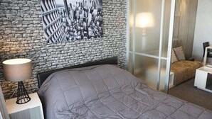 A bedroom with a stone wall, a bed with a grey quilt, a bedside lamp, and a view of a cityscape through a glass door. - D Condo Sign Chiang Mai R628 (Chiang Mai)
