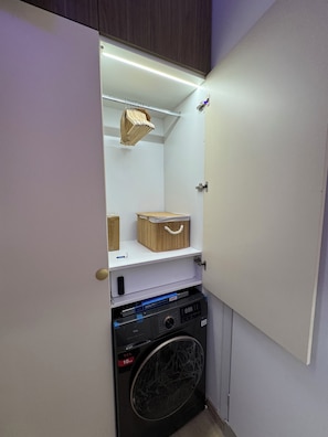 A compact laundry room with a washing machine, a drying rack, and a small shelf with a basket and a remote control. - Athens Green Smart Boutique Apartment near Syntagma and Ermou (Athina)