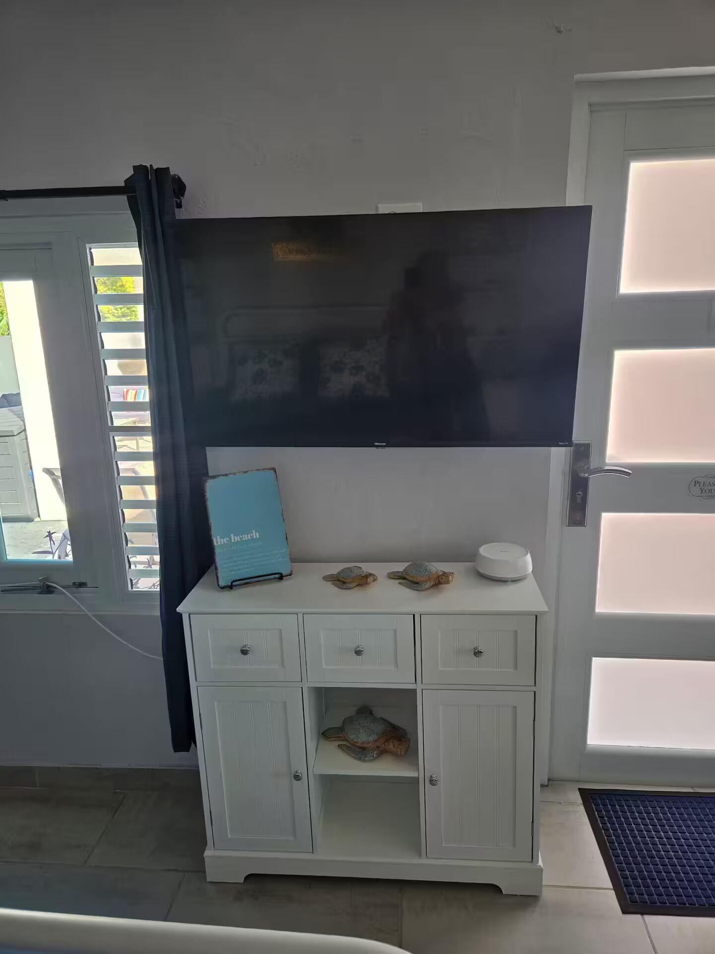 Una habitación con un armario blanco, un televisor de pantalla plana montado encima y una puerta con un letrero que dice 'Por favor, espere'.