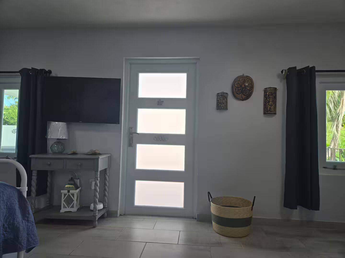 A room with a white door, a small white table with a lamp, a wicker basket, and a view of greenery outside.