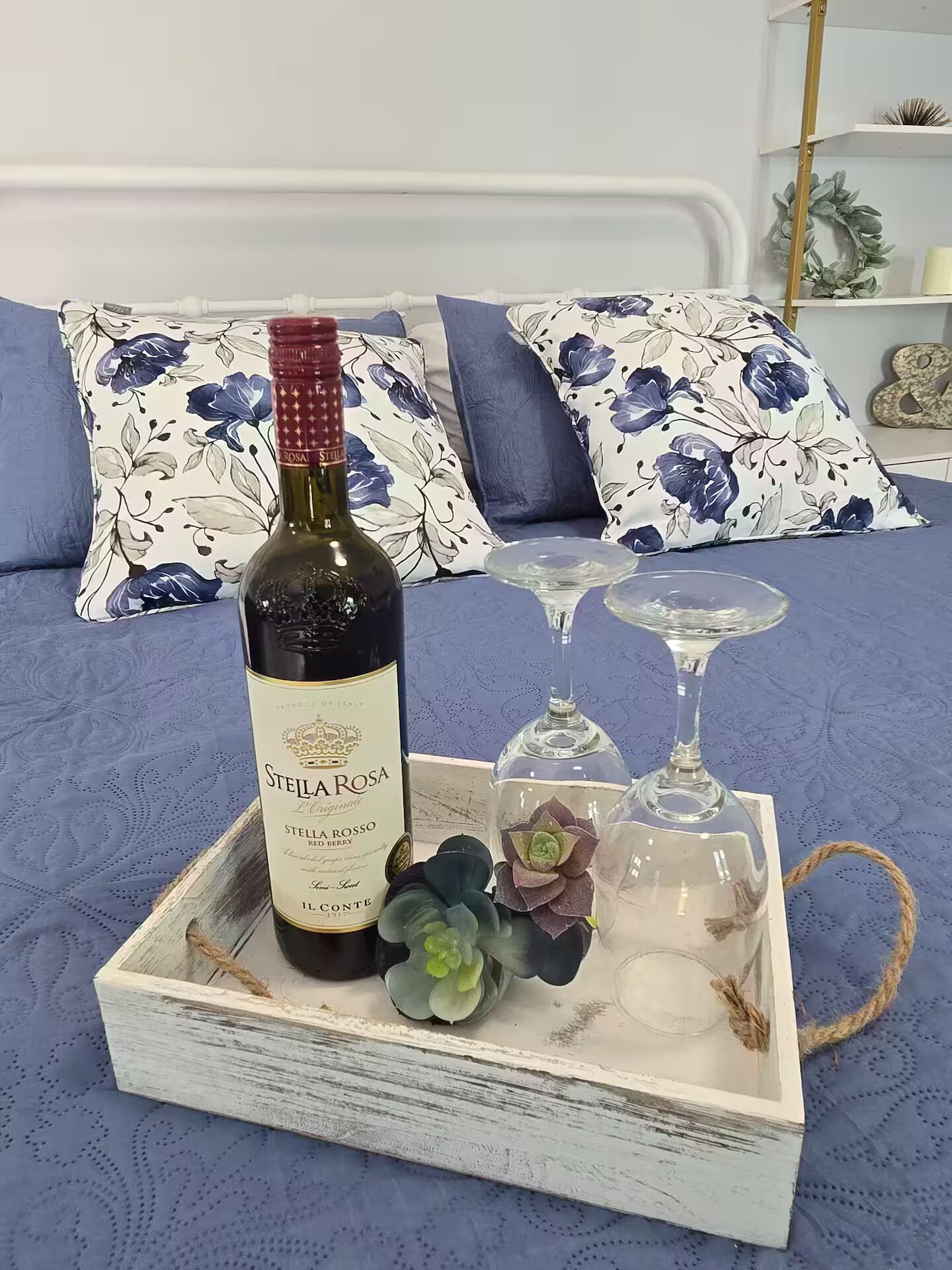 A wooden tray with a bottle of Steila Rosa red berry wine, two glasses, and decorative succulents on a bed with floral-patterned pillows.