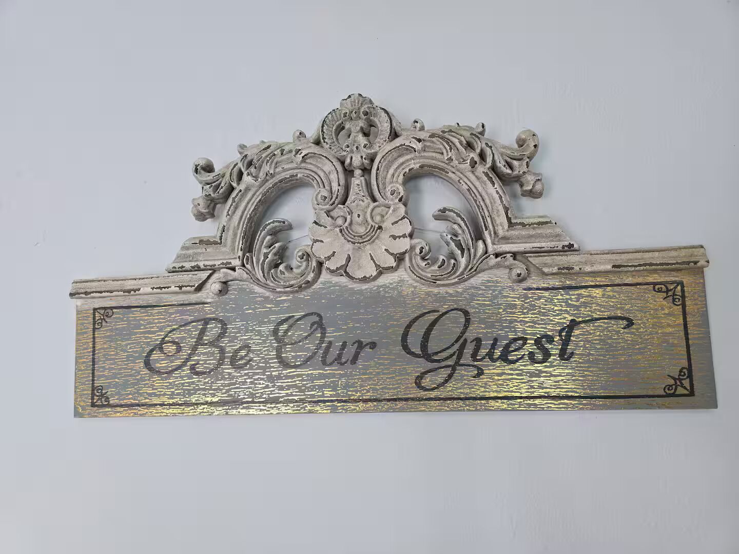 A decorative sign with the text "Be Our Guest" on a metallic background with ornate designs.