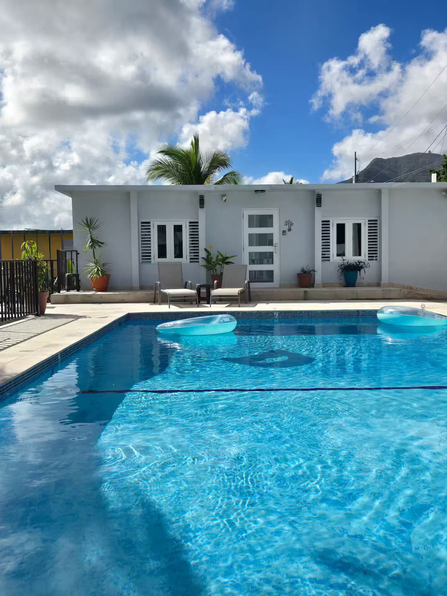A swimming pool with two inflatable rings, a white building with shutters, and potted plants.