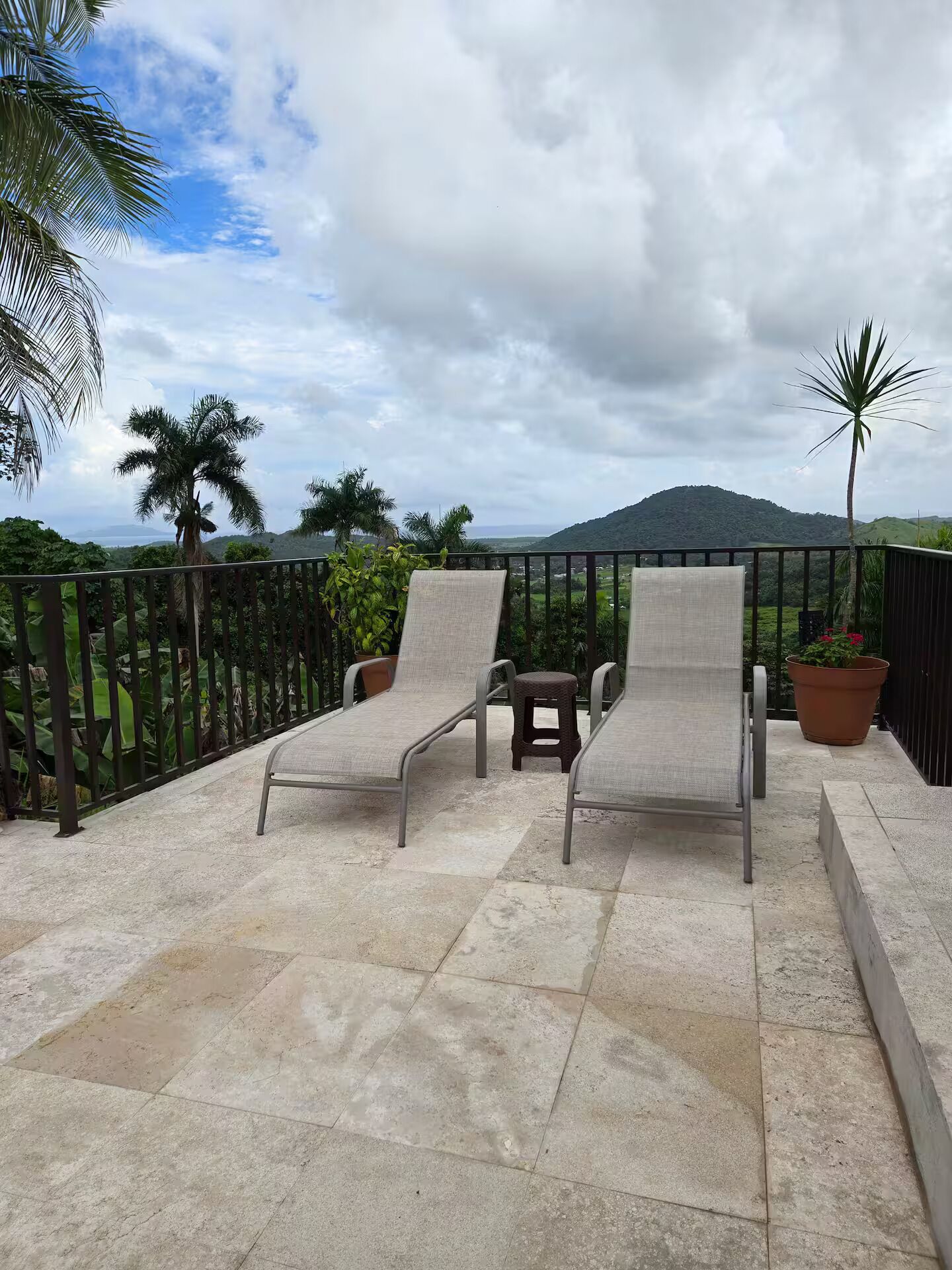 A balcony with two lounge chairs and a small table, surrounded by a metal railing, with a view of a mountainous landscape.