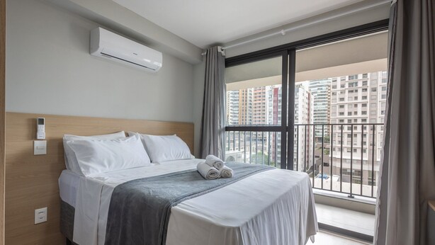 A hotel room with a bed, an air conditioning unit, a window with a view of high-rise buildings, and a control panel on the wall. - Complete Studios in Perdizes - CEP (São Paulo)