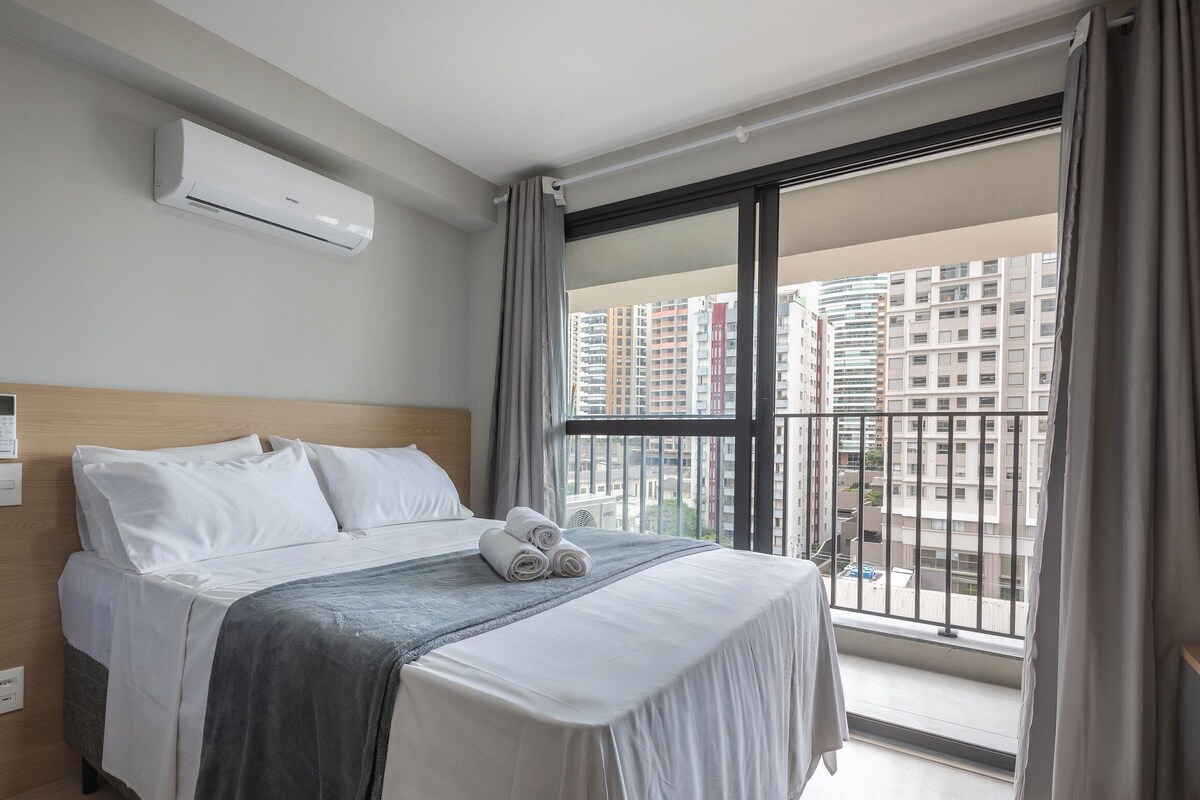 A hotel room with a bed, an air conditioning unit, a window with a view of high-rise buildings, and a control panel on the wall.