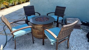 Een ronde tafel met een vuurkorf in het midden, omringd door vier rieten stoelen met blauwe kussens.