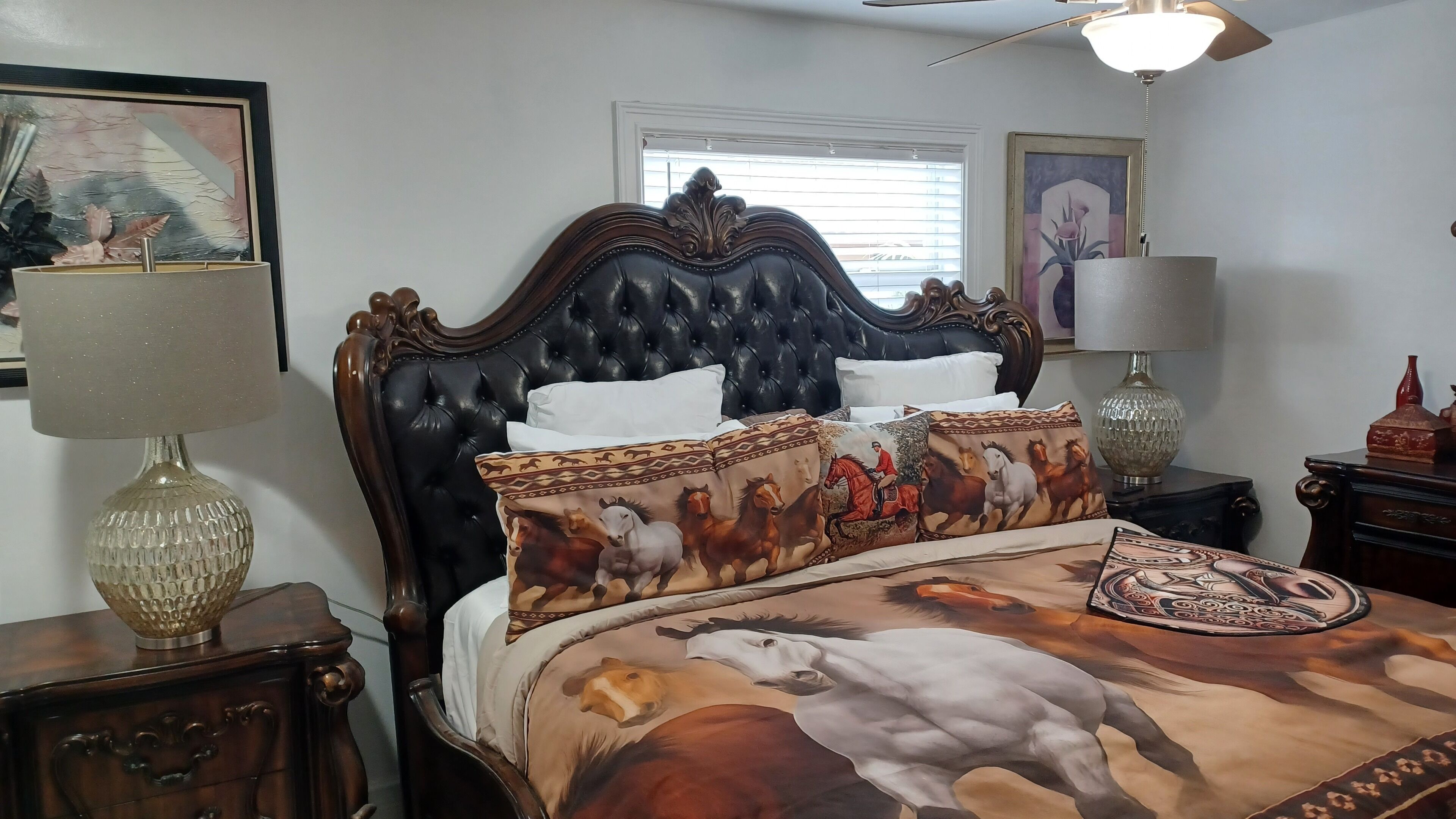 A bedroom with a dark upholstered headboard, a bed with a horse-themed bedspread, two bedside tables with lamps, and a window with blinds.