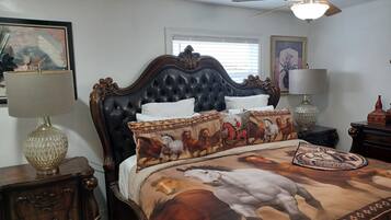 A bedroom with a dark upholstered headboard, a bed with a horse-themed bedspread, two bedside tables with lamps, and a window with blinds.