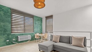Un salon moderne avec un canapé d’angle gris, une petite table ronde entourée de chaises et de grandes fenêtres équipées de stores.