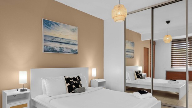 Une chambre d’hôtel moderne avec un grand lit, des tables de chevet, une petite table, un miroir et un tableau représentant une scène de plage.