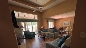 A spacious living room with a ceiling fan, a dining area with chairs and a table, and large windows allowing natural light. - Feels like Home on Hilton Head Island (Hilton Head Island)