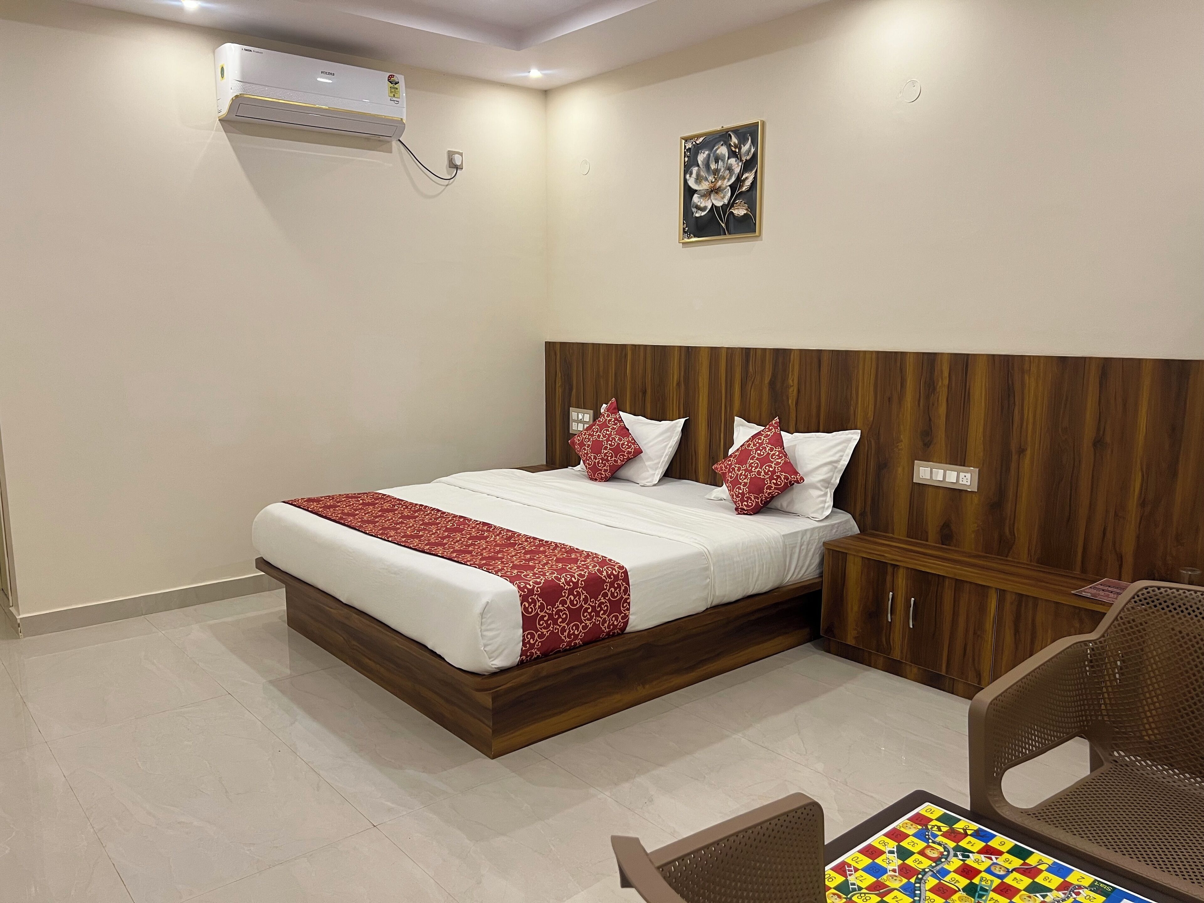 Una habitación de hotel con cabecera de madera, cama con ropa de cama blanca y almohadas con estampado rojo, un pequeño mueble de madera, una silla de mimbre y un juego de mesa en el suelo.